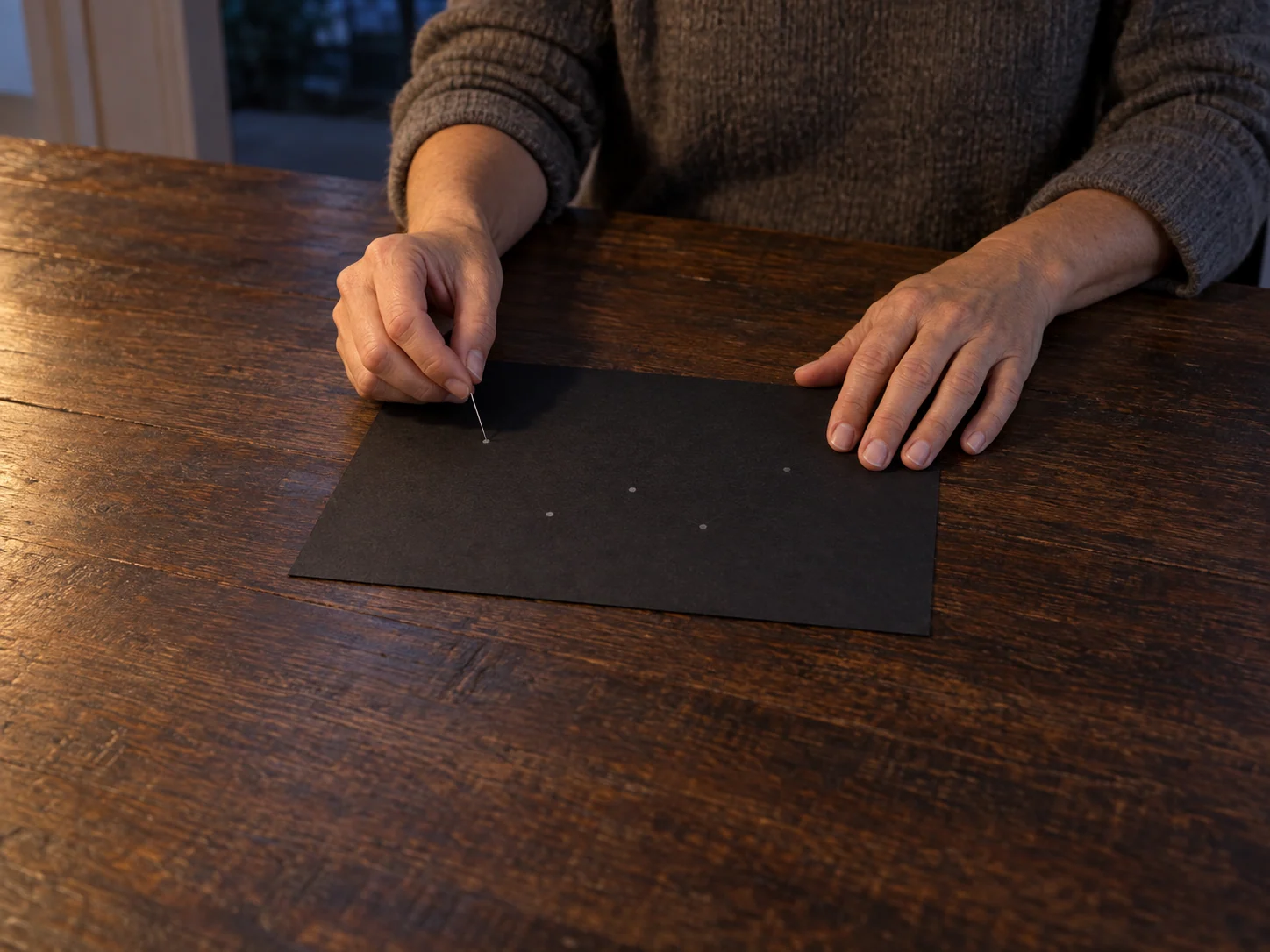 An adult woman’s hands place one pin at the first of five pencil marks on black paper.
